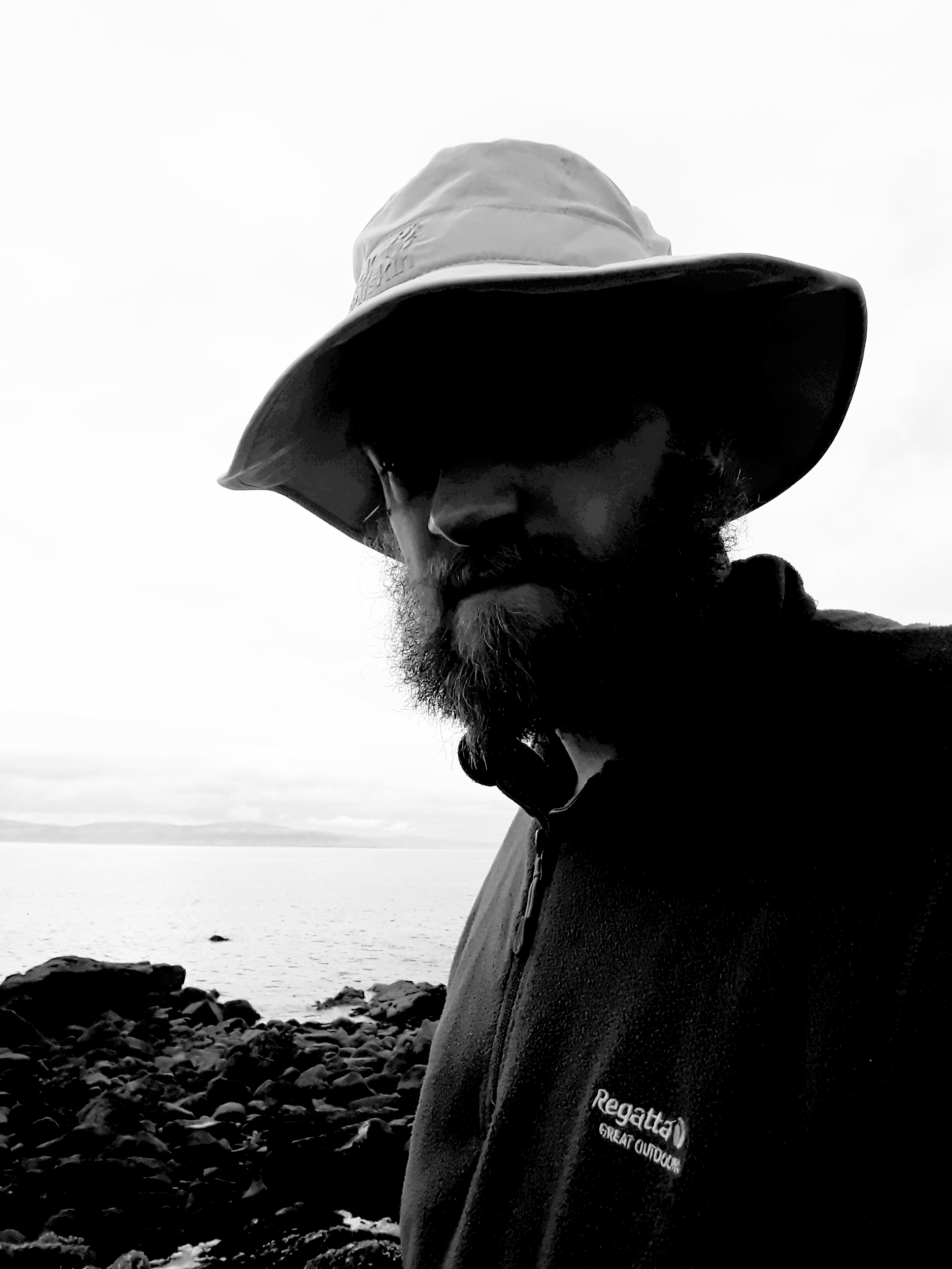 A black-and-white selfie of a man in a fleece and sun hat, framed against the sea